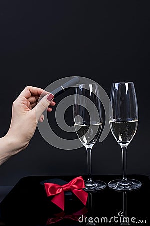 Black device in a female hand with red manicure and 2 glasses with champagne on a black background, Stock Photo