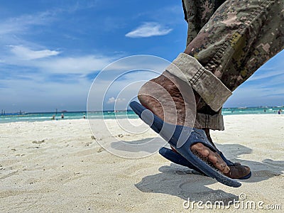 The black feet of an old man wearing blue sandals and military pants walking along the white sandy beach. Stock Photo