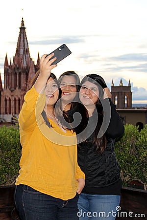 3 black haired latin women, sisters on outdoor terrace taking selfie with view of san miguel de allende parish in guanajoato mexi Stock Photo