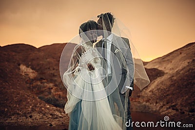 Newlyweds stand under bridal veil, smile and kiss in canyon at sunset. Stock Photo