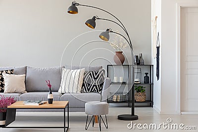 Black industrial lamp next to grey couch with patterned pillows, coffee table and pouf in monochromatic living room Stock Photo