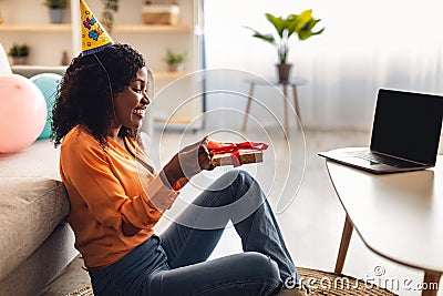 Black Lady At Laptop Opening Wrapped Birthday Present Indoors, Side-View Stock Photo