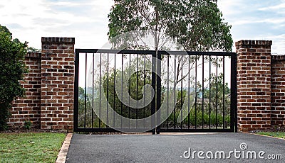 Black metal wrought iron driveway property entrance gates set in brick fence, lights, green grass, garden trees Stock Photo