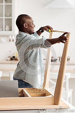 Black millennial handyman assembling wooden table and measuring the width between legs, standing in kitchen, crop Stock Photo