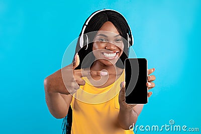 Black Millennial Woman Showing Phone Screen Wearing Headphones, Blue Background Stock Photo