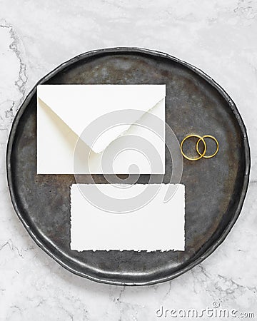 Black plate with a blank card, envelope and wto wedding rings top view, mockup Stock Photo