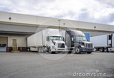 Black and white big rigs semi trucks with semi trailers loading cargo at warehouse dock with gates for each Stock Photo
