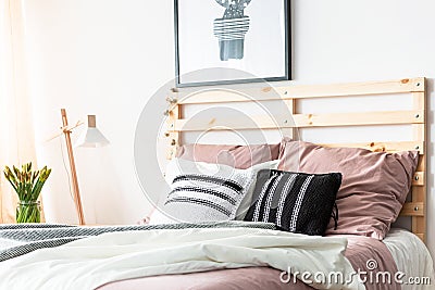 Black and white cushions placed on double bed with wooden bedhead in bright room interior with poster, fresh flowers and lamp Stock Photo