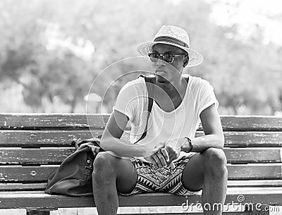 Black and White lifestyle fashion portrait. Stylish young african man sitting alone on a park bench wearing sunglasses and wicker Stock Photo
