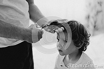 Black and white photo. Father cuts her son hair in the room. Family during quarantine, Scissors and a comb in male hands Stock Photo