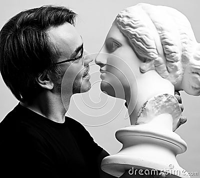 Black and white portrait of adult man in black t-shirt holding in hands antique sculpture woman head, going to kiss it Stock Photo