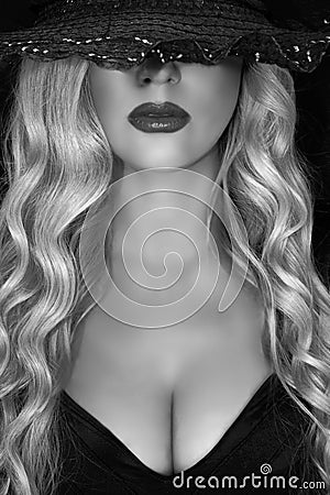 Black and white portrait of young blonde woman in black hat with black hat decollete and lush breasts, on a black background. Stock Photo