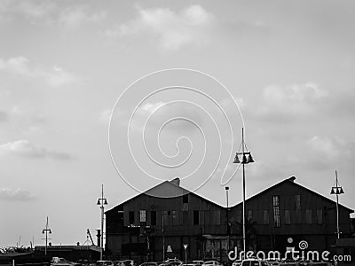 Black and white urban view of two roof tops of big buildings by the sea port, boat houses with tall city lamps in front, under Stock Photo