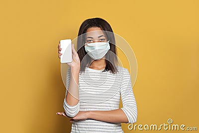 Black woman in medical face mask holding smart phone with empty display on vivid yellow background Stock Photo