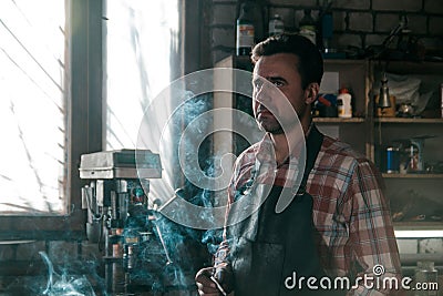 Blacksmith in leather apron smokes a cigarette at pause in work on the background of workspace Stock Photo