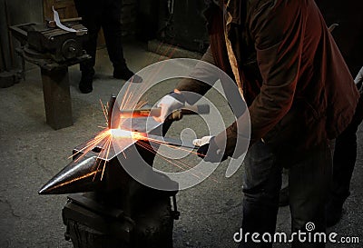 The blacksmith manually forging the molten metal on the anvil in smithy with spark fireworks. Spark. Dark background. Stock Photo