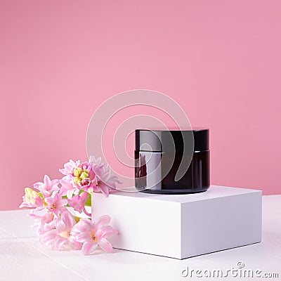 Blank amber glass cosmetic jar for cream on white podium and soft light pastel pink wall with fresh spring flowers. Mockup. Stock Photo