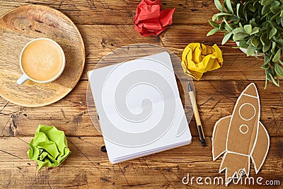 Blank notebook, coffee cup, plant and paper rocket on wooden office desk. Home workplace, freelancer and blogger concept Stock Photo