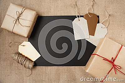 A blank sheet of black paper is surrounded by paper-wrapped gifts and gift tags on a canvas background. Stock Photo