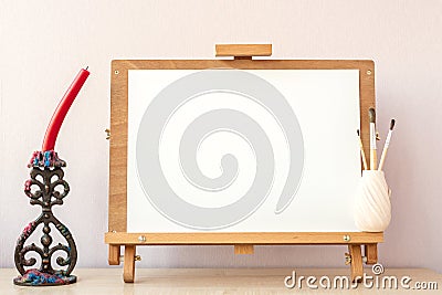 Blank white paper for painting on wooden easel, brushes and candle on table at home studio Stock Photo