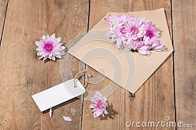 Blank white paper tag with brown envelope and pink flowers on wooden table. Stock Photo