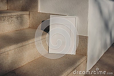 Blank white picture frame leaning against white wall. Outdoor sandstone stairs in sunlight, shadows overlay. Empty Stock Photo