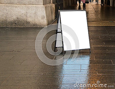 Blank white sandwich sign outside in daytime on dark wet sidewalk near a building. Stock Photo