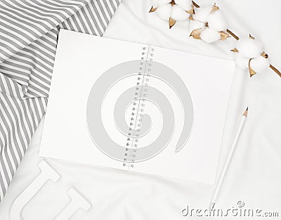 Blank white spiral notebook with pencil, cotton flowers, grey stripe fabric and wooden letter on white bed sheet. Stock Photo