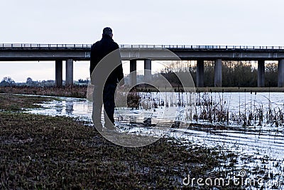 A bleak, moody, winter edit of a figure standing next to a lake, looking out to a motorway bridge, out of focus in the background Stock Photo