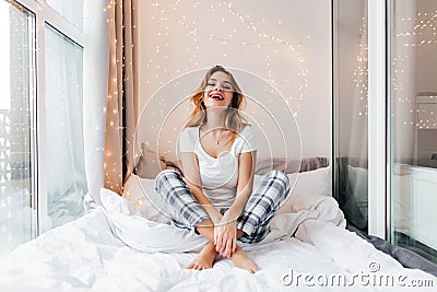 Blithesome girl in white t-shirt laughing during photoshoot at loggia. Indoor shot of sensual blonde lady resting in bed Stock Photo