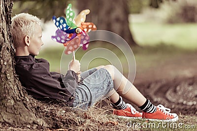 Blond boy holding colorful whirligig sits by tree Stock Photo