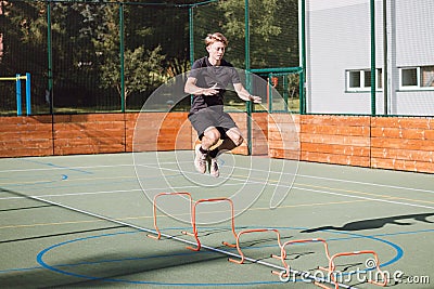 Blond boy in sportswear jumps over red obstacles to improve lower body dynamics. Plyometric training in an outdoor environment. Stock Photo