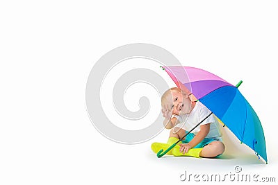 A blond child caucasian sits under a rainbow bright umbrella in rubber boots and waves his hand on a white background Stock Photo