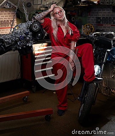 Blond female mechanic with tattooed hands in orange overalls stands in garage or workshop Stock Photo