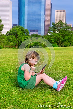 Blond kid girl playing with smartphone sitting on park lawn at c Stock Photo