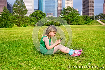 Blond kid girl playing with smartphone sitting on park lawn at c Stock Photo