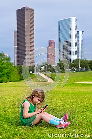 Blond kid girl playing with smartphone sitting on park lawn at c Stock Photo