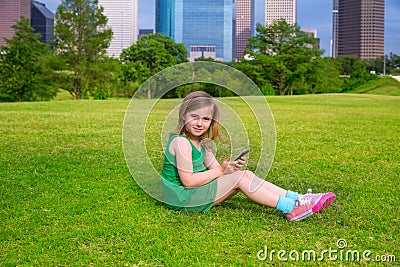 Blond kid girl playing with smartphone sitting on park lawn at c Stock Photo