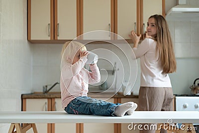 Blond little girl drinking from a large mug. Mom looks after the baby in the kitchen Stock Photo