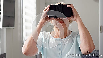 Blond senior woman in white using VR 360 glasses at home. Concept of active modern elderly people Stock Photo