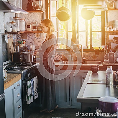 Blond tired woman making coffee in an old narrow cluttered kitchen at sunset Stock Photo