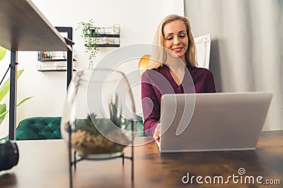 Blond white woman wearing maroon shirt sitting by desk in living room working on laptop and smiling. Horizontal indoor Stock Photo