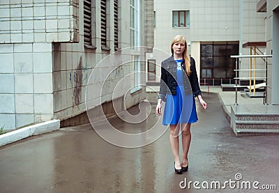 Blond woman in blue dress and denim jacket walking down the city street against background of urban architecture Stock Photo