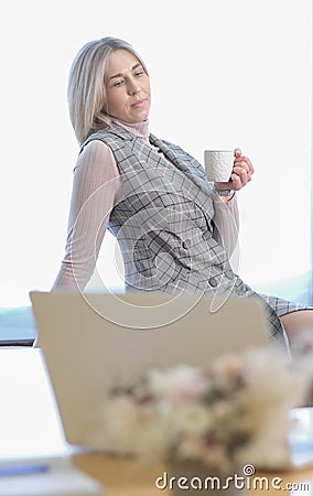 blond woman wearing office clothes sitting on a windowsill with cup of coffee getting rest. coffee break, hard working Stock Photo