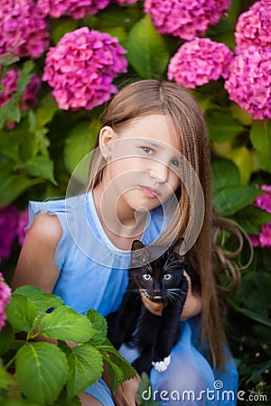 A blonde girl in a blue dress holds a black cat with white spots in her arms, a girl in the garden Stock Photo