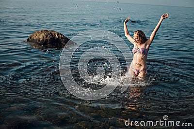 A blonde girl with long hair, in a striped swimsuit, bathes in the ocean, laughs, plays and splashes Stock Photo