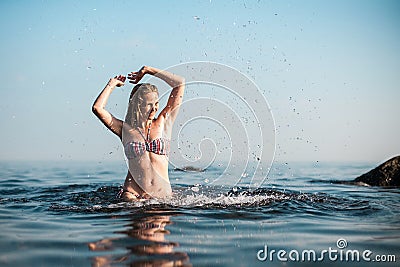 A blonde girl with long hair, in a striped swimsuit, bathes in the ocean, laughs, plays and splashes Stock Photo