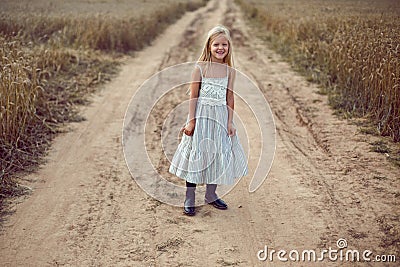 Blonde girl stands on an earthen path in a long dress among a wheat field Stock Photo