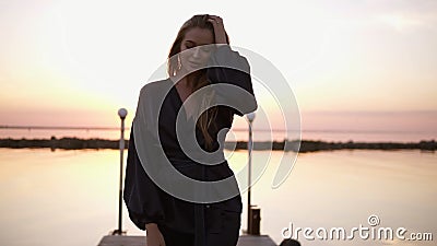 Blonde haired young caucasian woman, model in black clothes kimono walking by the pier. Morning pink light. Seaside stock footage Blonde haired young caucasian woman, model in black clothes kimono walking by the pier. Morning pink light. Seaside stock footage