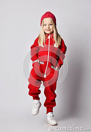 Blonde kid girl in white sneakers, red coldproof jumpsuit and winter hat stands lifting foot to step over, walking Stock Photo